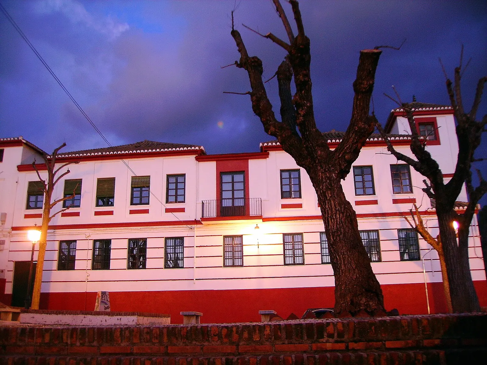 The red and white facade of the former Hospital Militar on the Campo del Príncipe at dusk, with bare trees under a violet sky