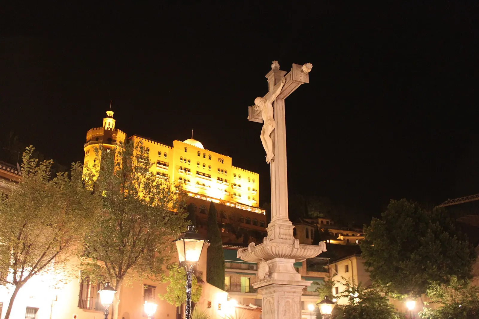The stone cross of the Cristo de los Favores lit at night in the Campo del Príncipe, with street lamps, trees and lit buildings on the hill beyond
