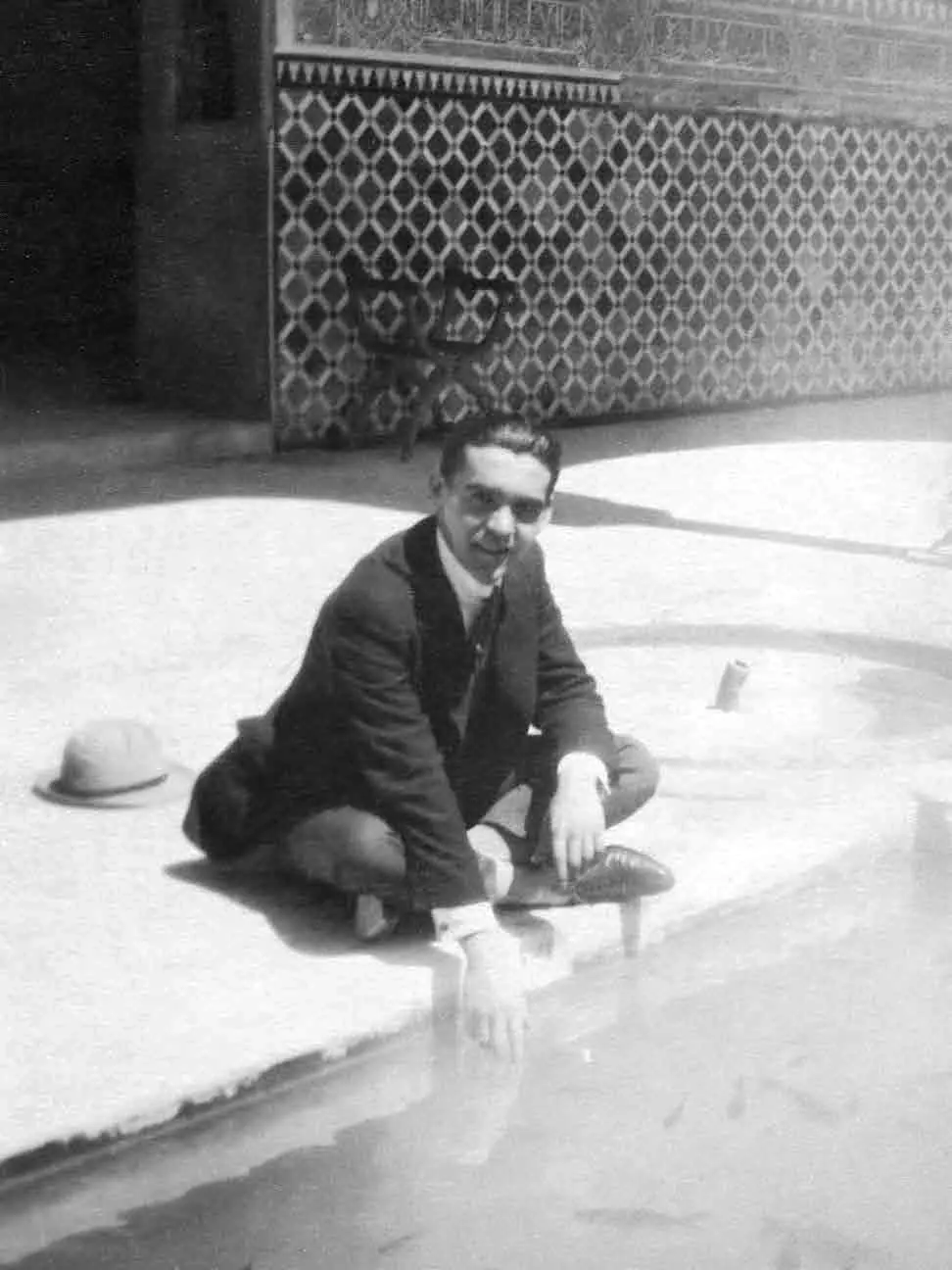 A black and white photograph of Federico García Lorca sitting at the edge of a pool in an Alhambra courtyard, with tilework behind him