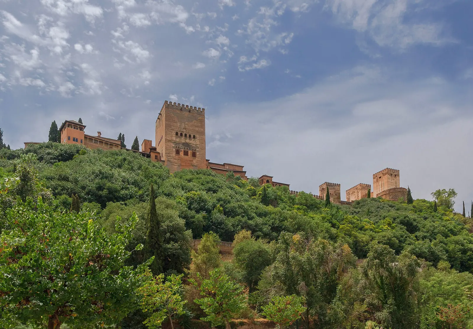 The Alhambra on its wooded hill seen from below the Paseo de los Tristes, under a partly cloudy sky